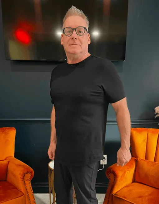 Man with short gray hair and glasses wearing a black t-shirt and pants standing between two orange armchairs in a room.