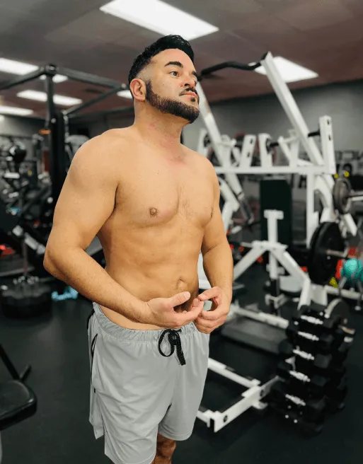 Shirtless man with a beard wearing gray shorts standing in a gym with weightlifting equipment in the background.