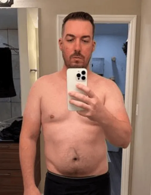 Shirtless man with short hair taking a selfie in a bathroom mirror using a smartphone.