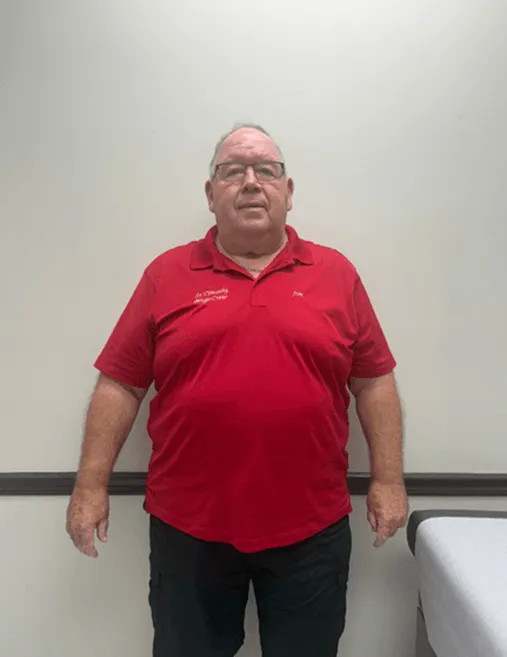 Man wearing glasses and a red polo shirt standing against a plain white wall next to a white table.