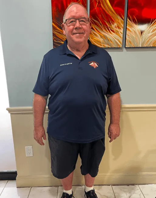 Older man wearing glasses, a navy blue polo shirt, black shorts, white socks, and black shoes standing indoors in front of a beige wall with three red and orange decorative panels.
