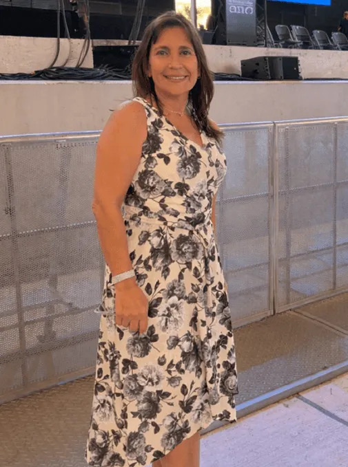 Smiling woman standing indoors wearing a white dress with black floral patterns.