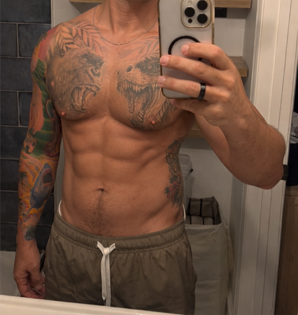 Man taking a mirror selfie wearing a sleeveless navy dress with white trim and white shoes in a room with dark flooring and white walls.