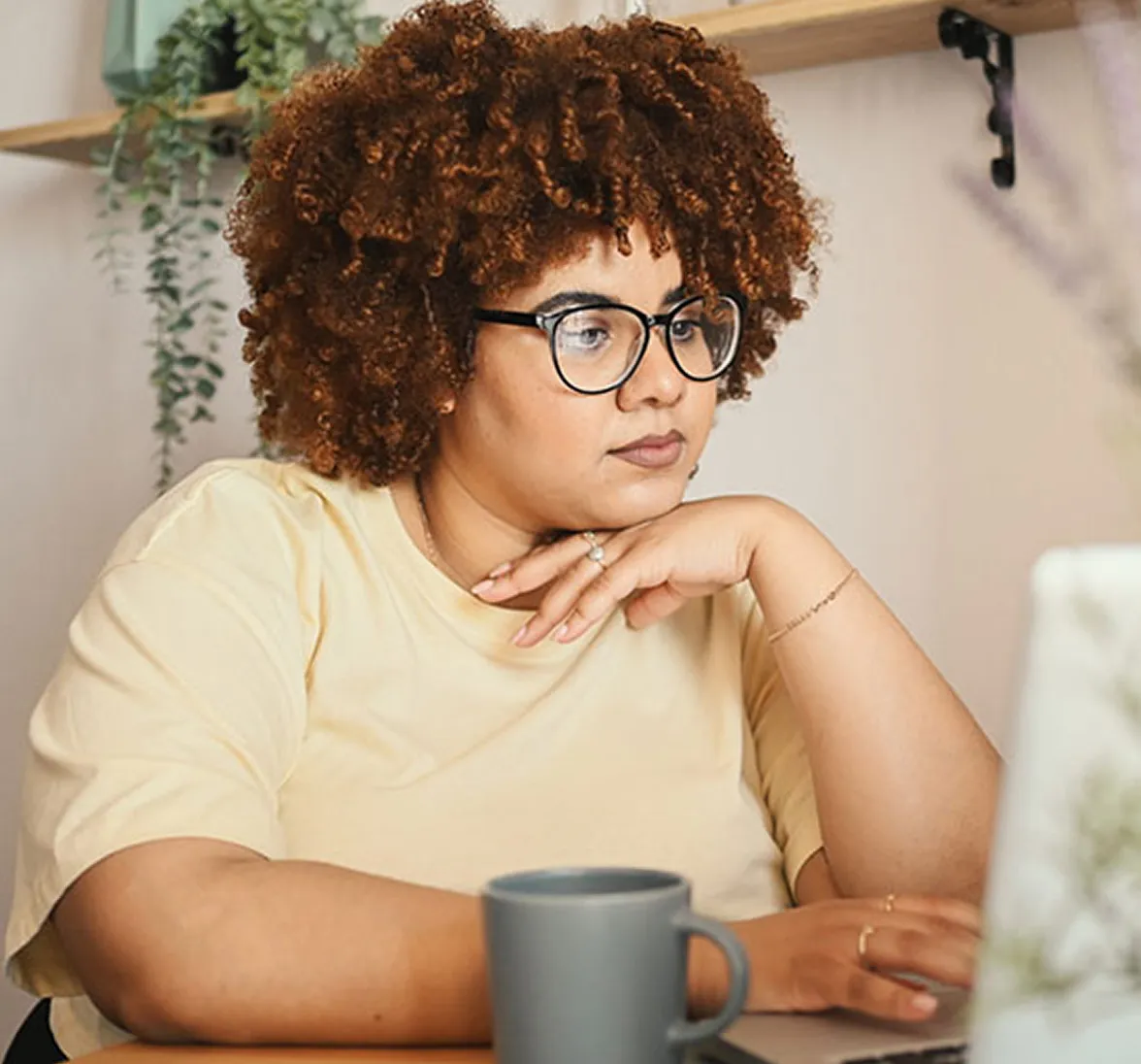 Focused woman with curly hair and glasses working on a laptop at home with a coffee mug nearby.