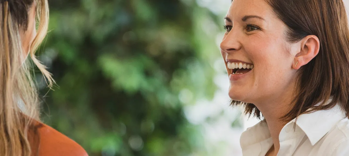 Smiling woman engaged in a cheerful conversation with a colleague in a natural, green setting.