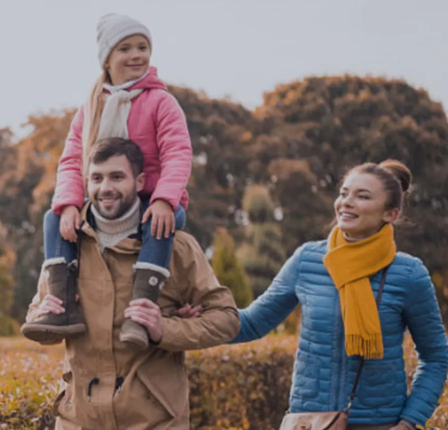 Smiling family walking outdoors in autumn, with child on father's shoulders and trees in background.