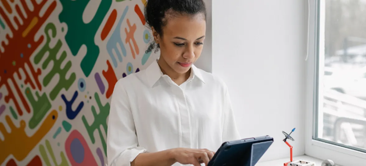 Woman in white blouse using a tablet near a window with colorful abstract wall art.