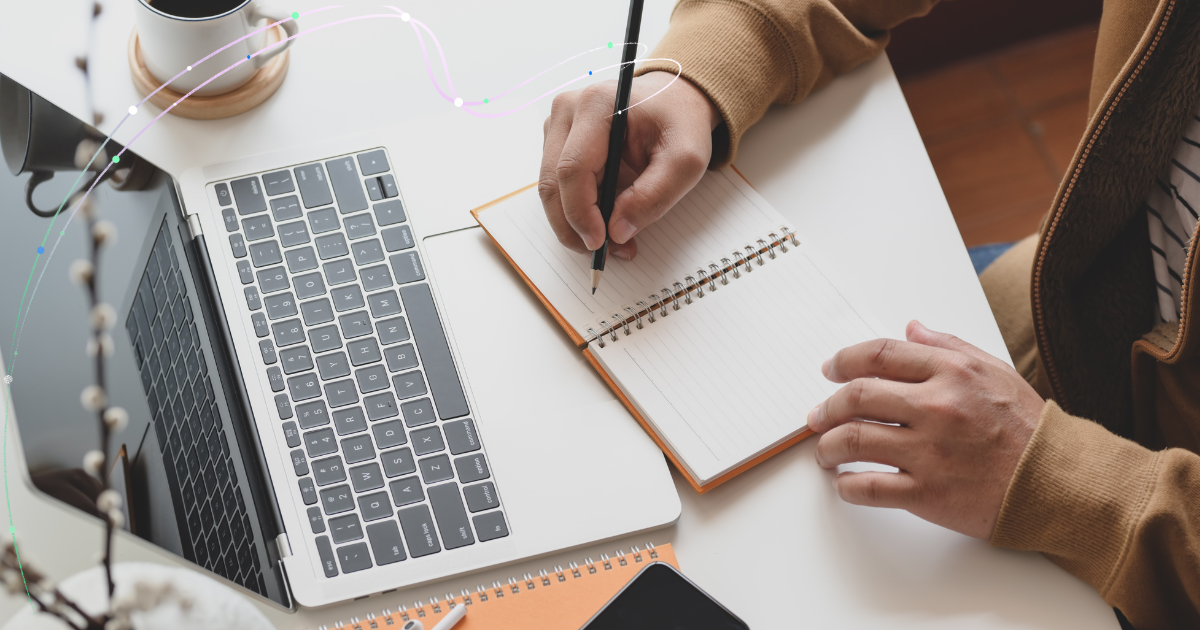 Top view of a person typing on a laptop at a white desk, with a smartwatch on their wrist. Nearby are a plant on a notebook, eyeglasses, two pencils, and a marble-patterned phone case.