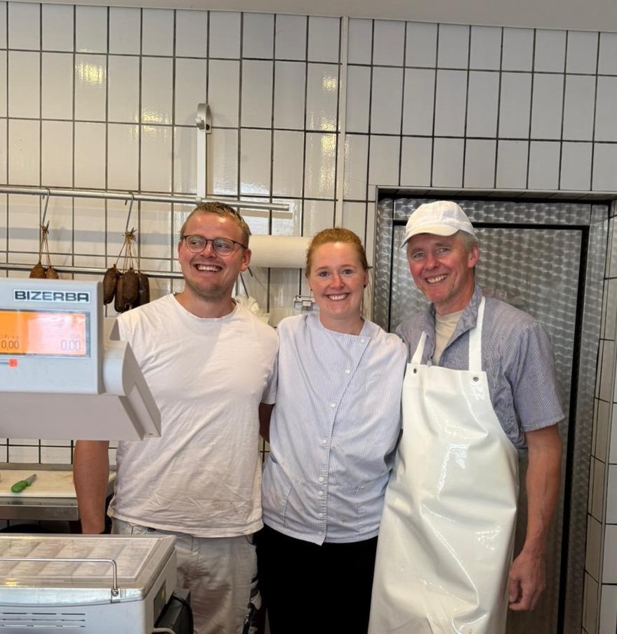 Ein Mann in formeller Kleidung hält stolz seinen Meisterbrief in der Hand. Im Hintergrund sind Kühe auf einer Wiese zu sehen, umgeben von einem Zaun. Der Standort deutet auf einen ländlichen Hofladen hin.