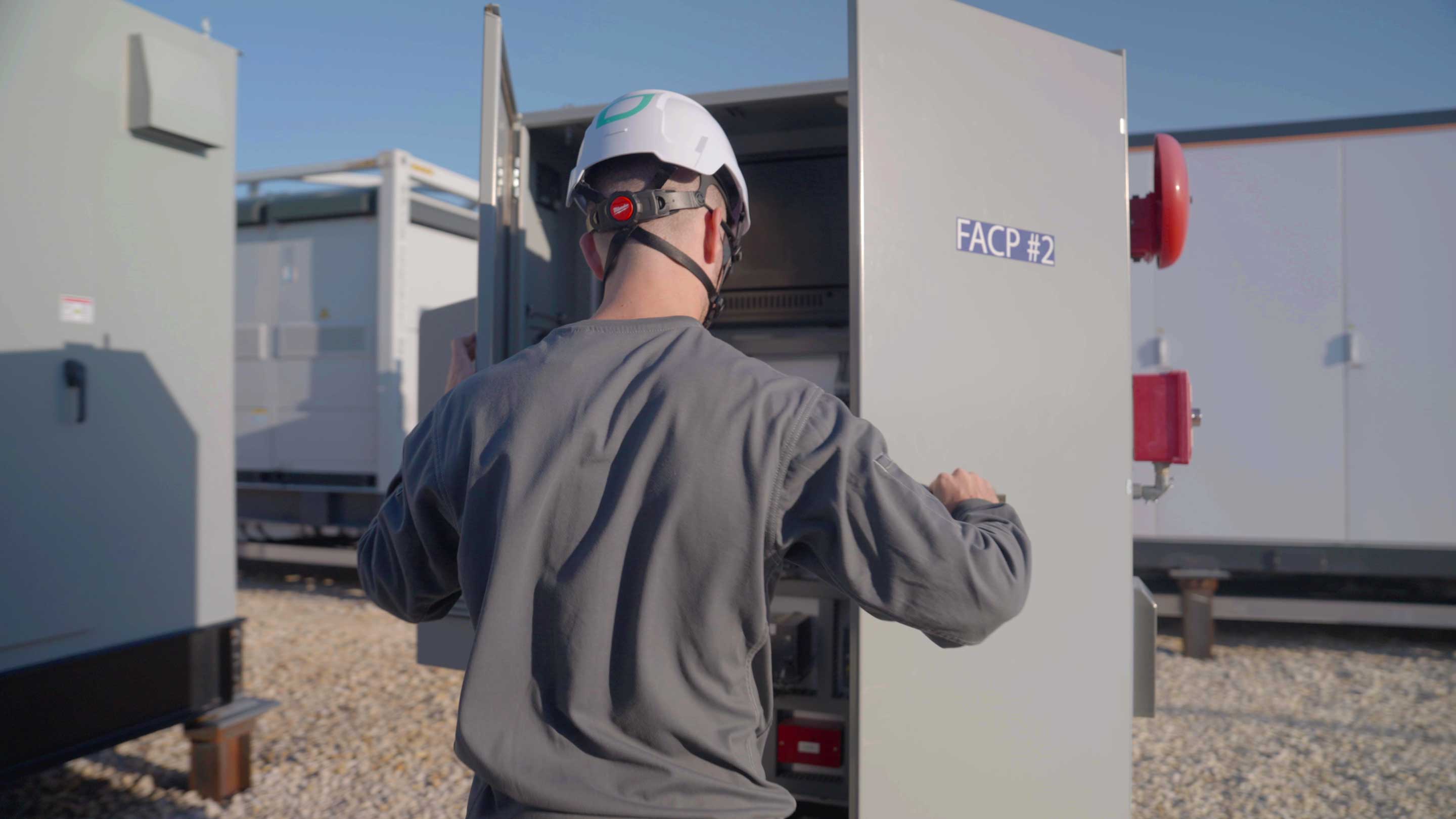 Worker in helmet and gray shirt opening a large metal electrical cabinet labeled FACP #2 outdoors.