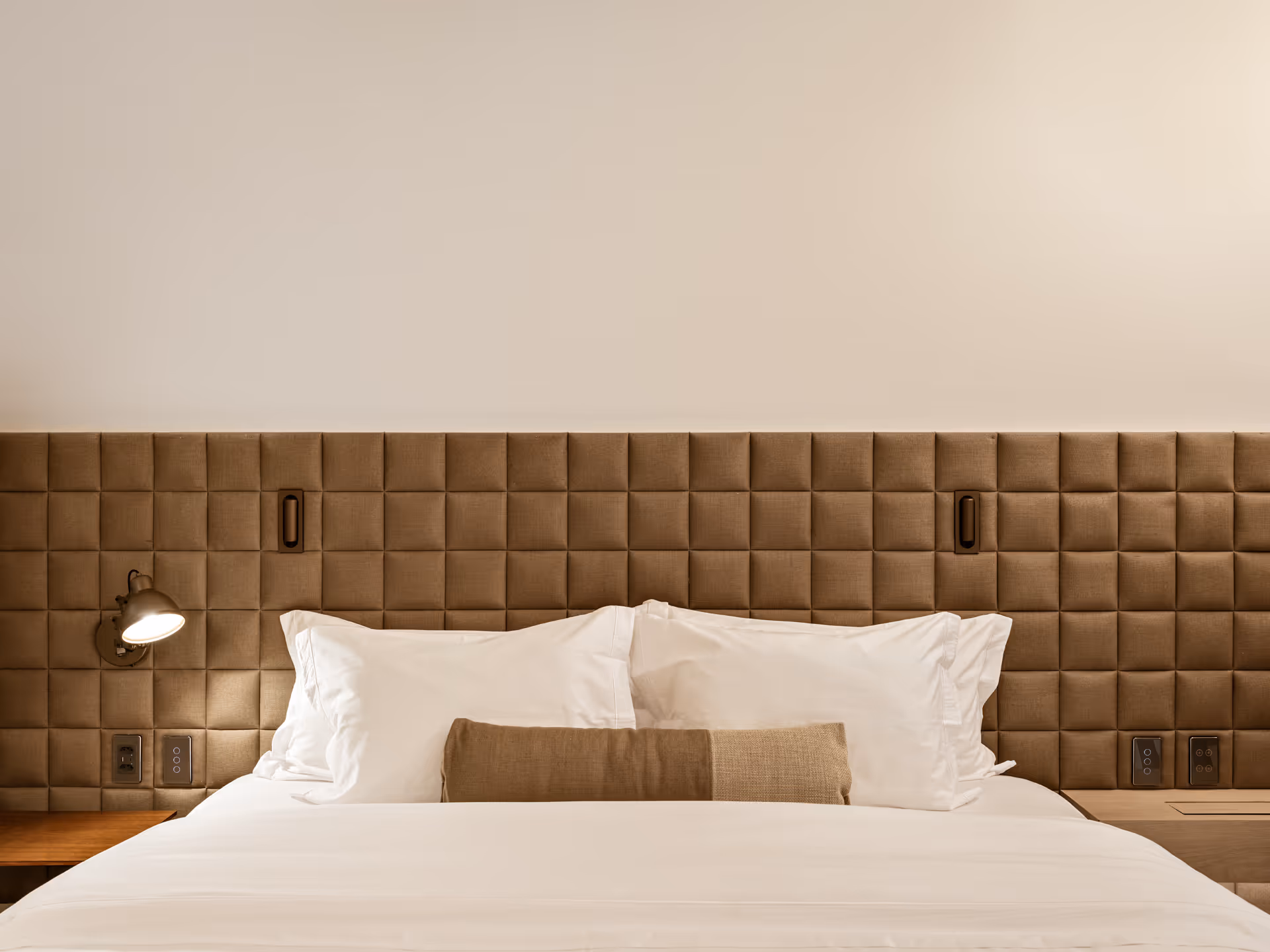 A modern bed with a tufted beige headboard, two white pillows, and a decorative pillow in the center. Wall-mounted lamps are attached to either side of the headboard.