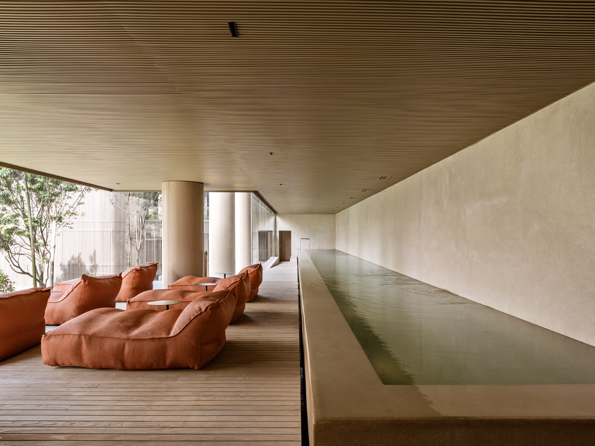 An indoor seating area featuring large, orange bean bag chairs arranged on wooden flooring, alongside a narrow water feature against a smooth white wall.