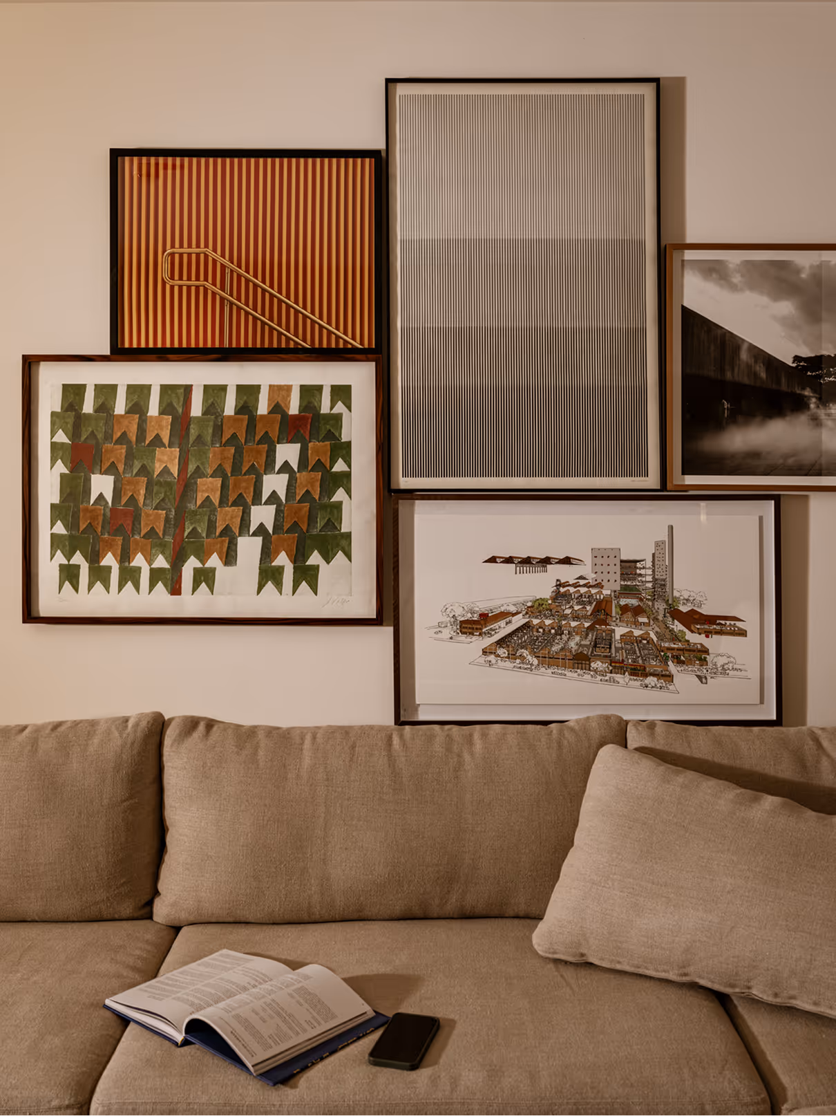 A cozy living room scene featuring a beige couch with an open book and a smartphone on it. On the wall behind, several framed artworks are displayed in varying styles and colors, including abstract designs and a landscape illustration.