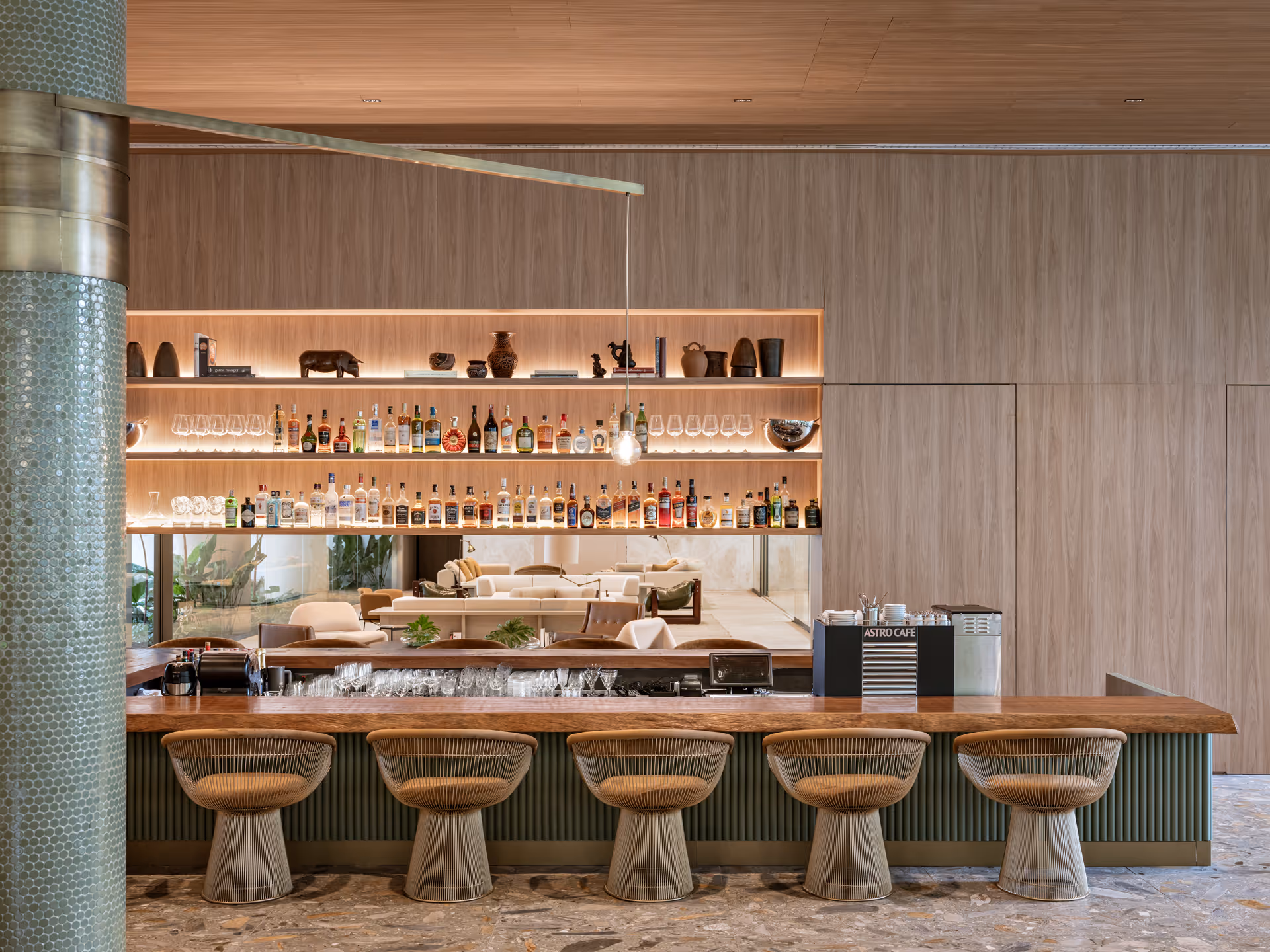 A stylish bar area featuring a wooden counter with round stone stools. Behind the bar, a wall of shelves displays various bottles and glassware, illuminated by soft lighting. A contemporary seating area is visible in the background.