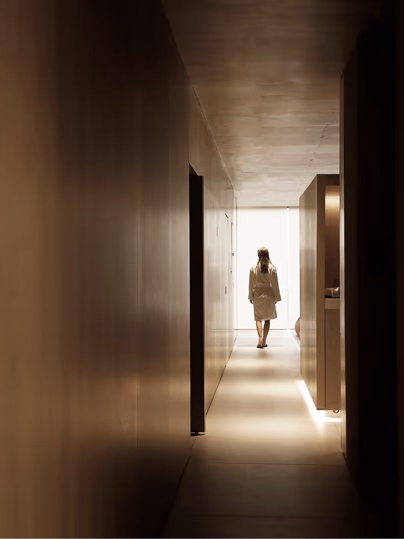 A person wearing a white robe walking away down a hallway with soft lighting, surrounded by minimalistic walls and doors.