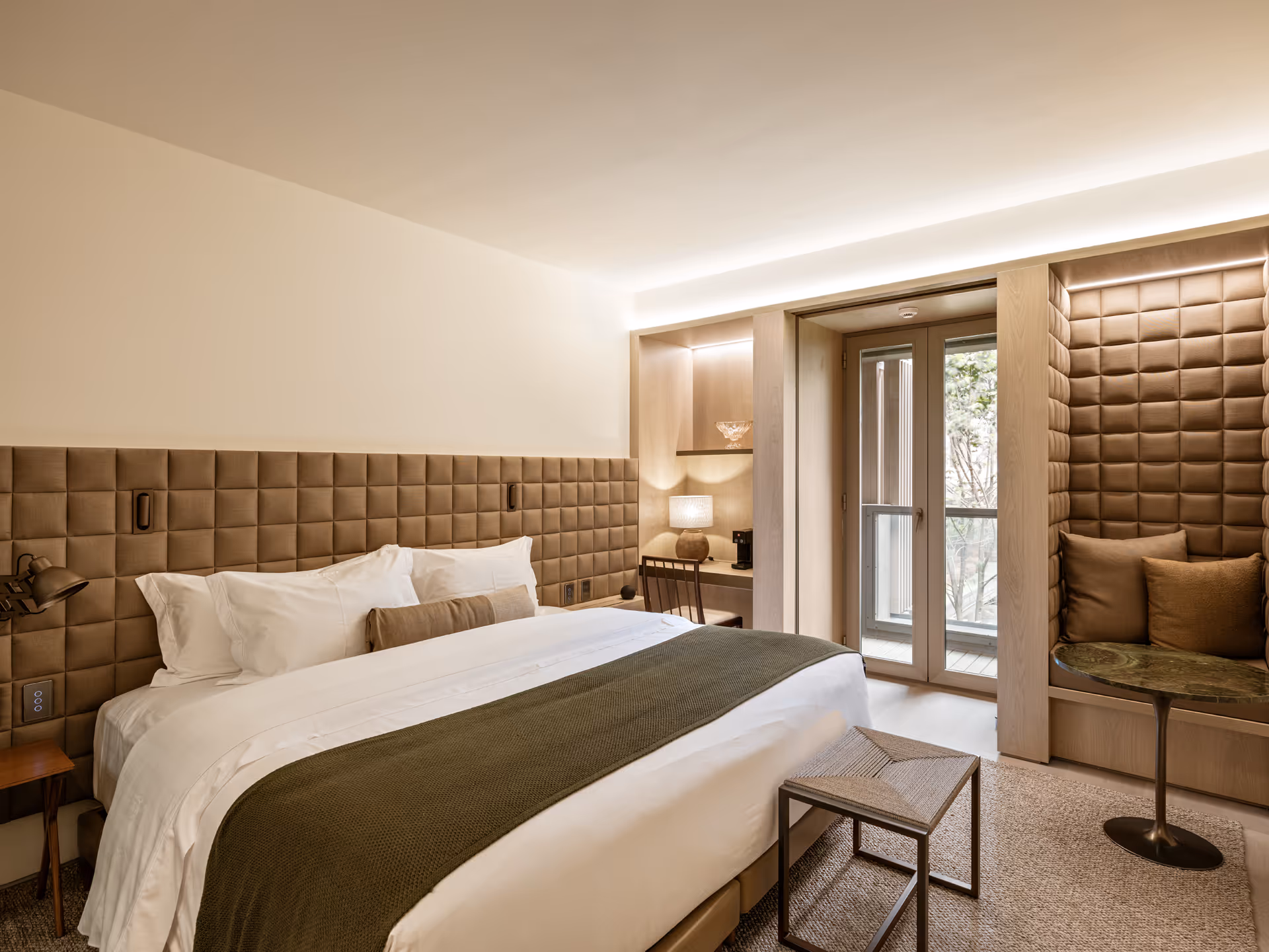 A modern hotel room featuring a king-sized bed with a brown throw blanket, plush headboard, and a small table. Natural light filters through a glass door leading to a balcony, with a cozy seating area by the window.