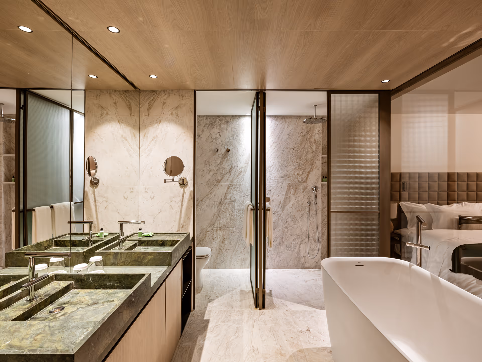 A modern bathroom featuring dual sinks with green marble countertops, a glass shower enclosure, and a free-standing bathtub. Soft lighting highlights the sleek design with wooden elements and gray tiles.
