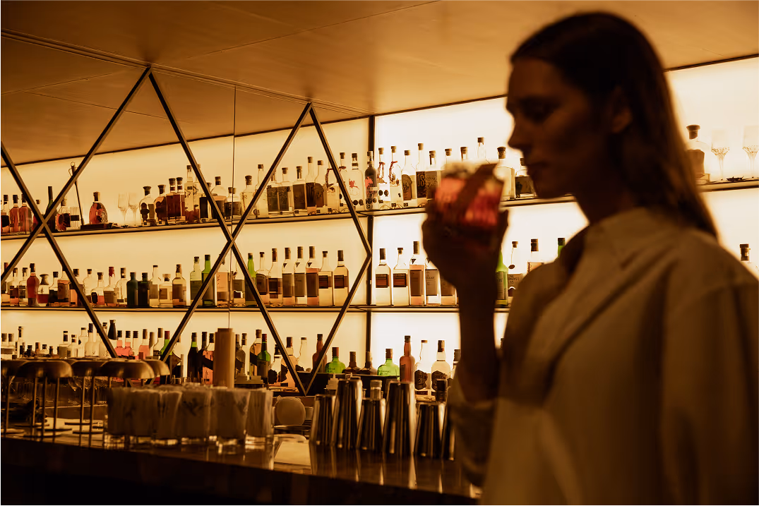 Woman sipping a pink cocktail in an upscale modern bar with extensive drink menu.