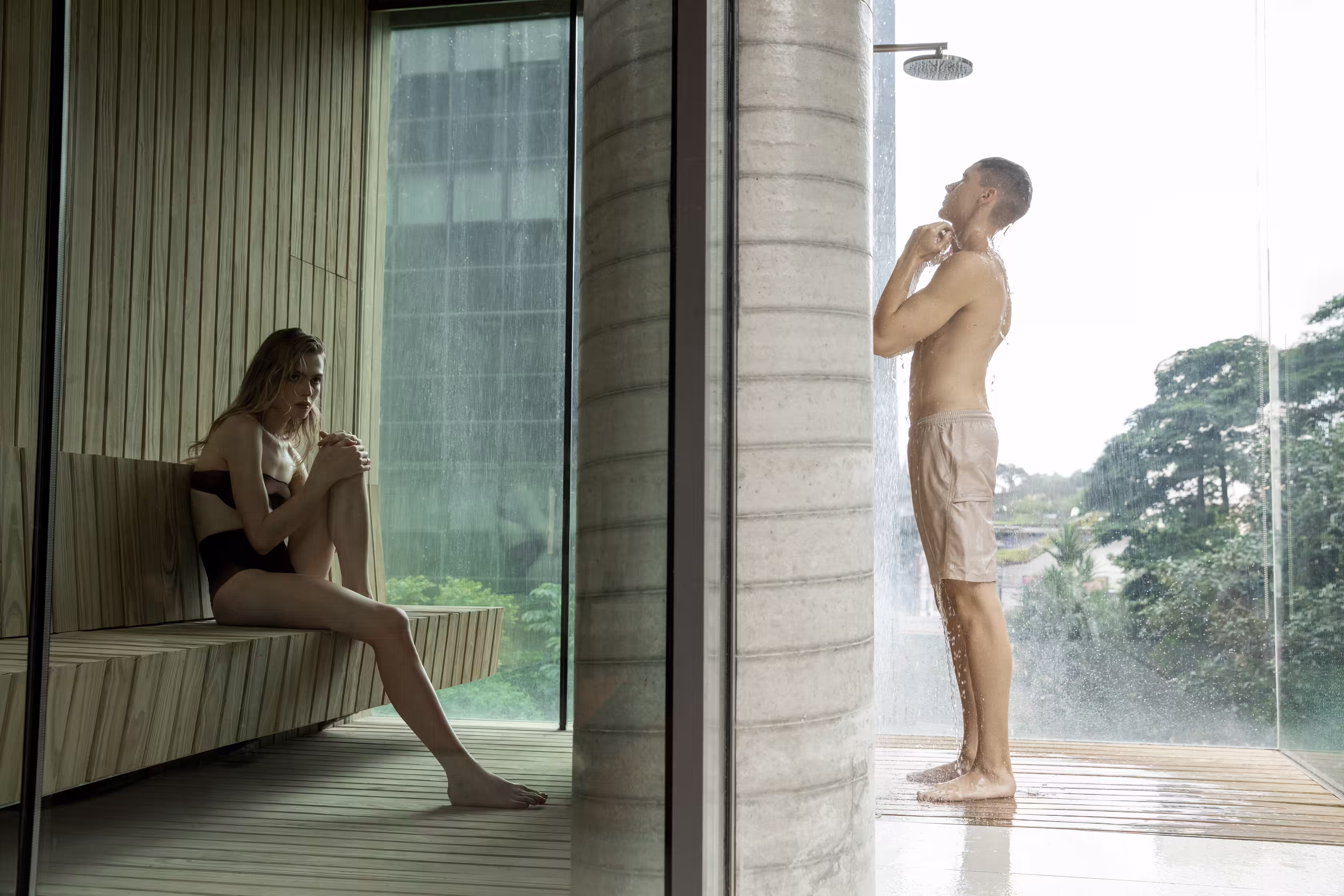 A modern sauna with a wooden interior. A woman lies on a wooden bench, and a man stands nearby, both in relaxed poses. Large glass windows show a view of greenery outside.