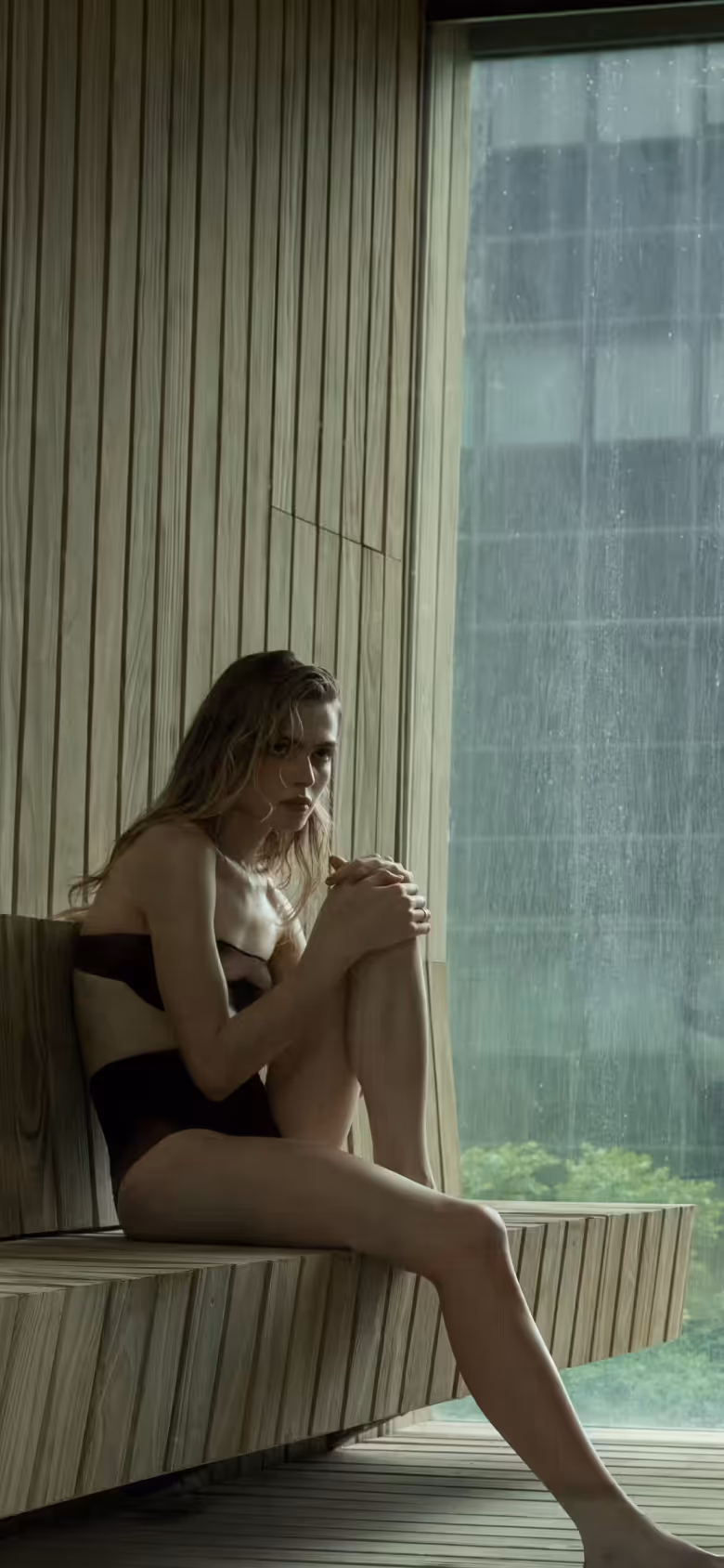 A modern sauna with a wooden interior. A woman lies on a wooden bench, and a man stands nearby, both in relaxed poses. Large glass windows show a view of greenery outside.