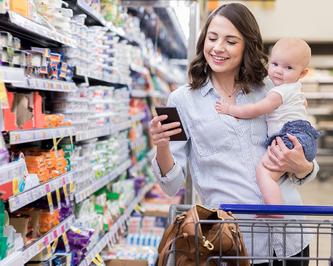 A woman holding her baby while we is looking at her phone at a grocery store. 