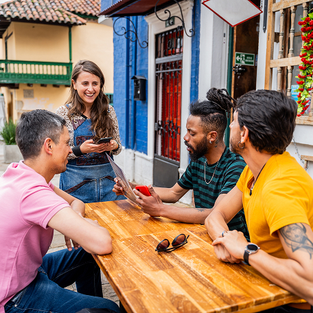 Group of friends sitting at a wooden table outdoors, placing an order with a smiling waitress.