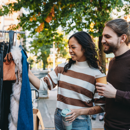 Smiling couple browsing clothes at an outdoor market while holding coffee cups.