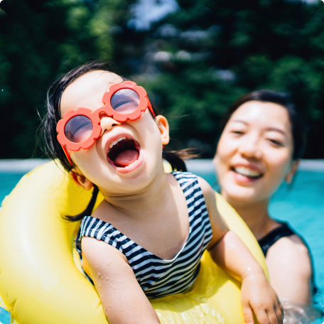 Young child in bright sunglasses laughing in a yellow pool float with parent.