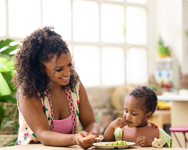 Mother helping her toddler eat a healthy meal at the dining table.
