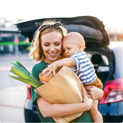 Smiling mother holding her baby and a paper bag of groceries near the car.