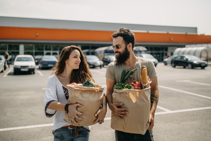 A smiling man and woman carrying paper grocery bags filled with fresh vegetables, fruits, and bread walk together in a parking lot outside a supermarket on a sunny day.