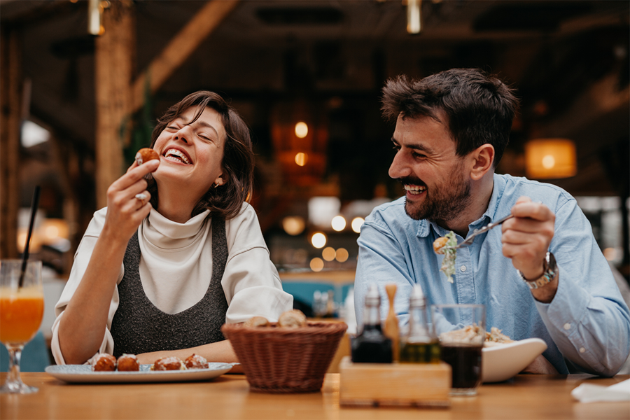 A cheerful couple sitting at a restaurant table enjoying a meal together; the woman laughs while holding a bite of food, and the man smiles as he eats. The setting is warm and cozy with ambient lighting.