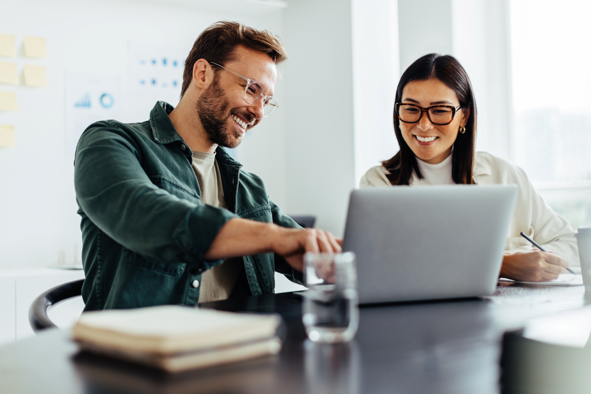Two colleagues smiling and working together on a laptop during a meeting in a modern office.