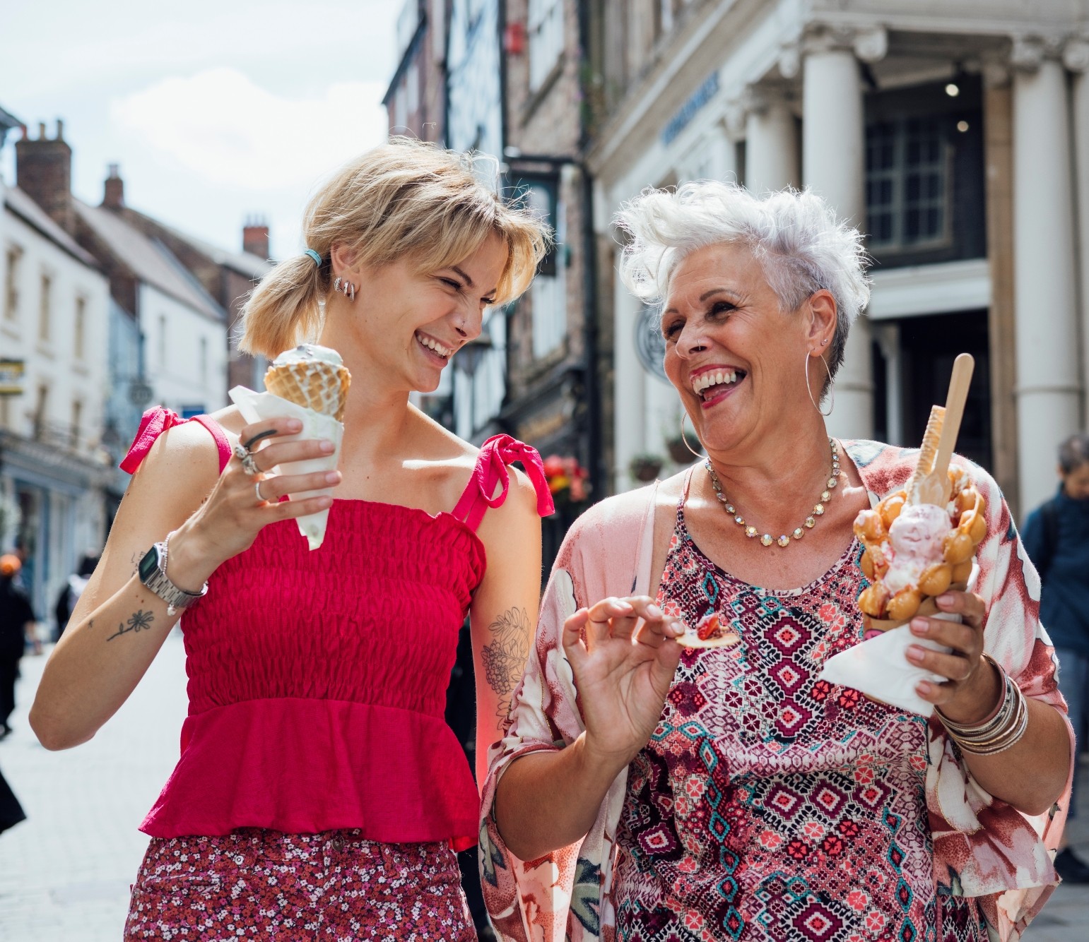 Two women smiling and enjoying ice cream together while walking down a sunny city street.