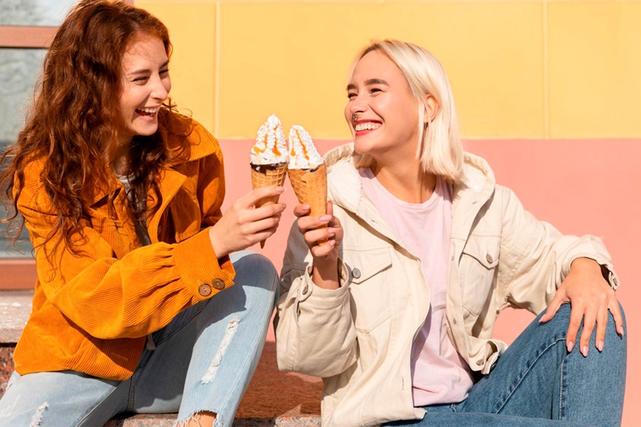 Two friends sitting outdoors in the sunshine, smiling and clinking their ice cream cones together in front of a colorful wall.