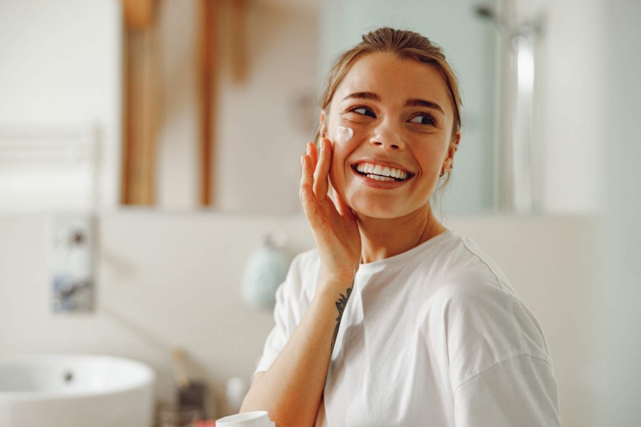 A smiling woman applying face cream in a bright bathroom, looking off to the side while performing her skincare routine.