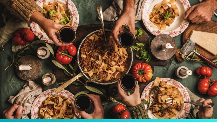 Top-down view of a shared pasta dinner, with several hands reaching for plates and holding glasses of wine around a rustic table.