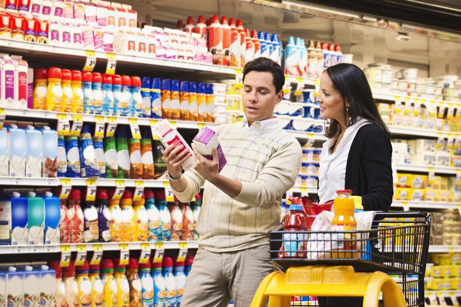 A man and woman standing in a supermarket aisle, comparing two cartons of refrigerated products while their shopping cart sits nearby.