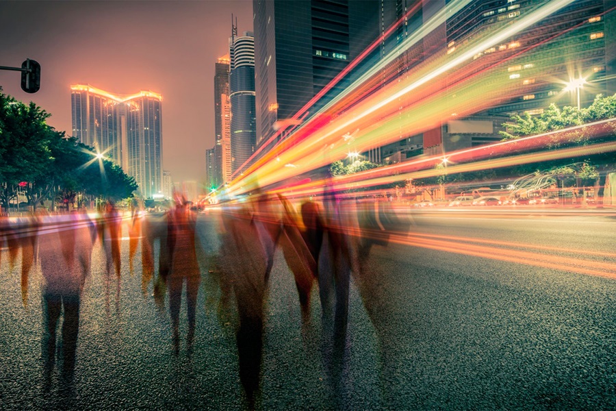 Blurred silhouettes of people walking on a city street at night, with colorful light trails from passing vehicles creating dynamic motion.
