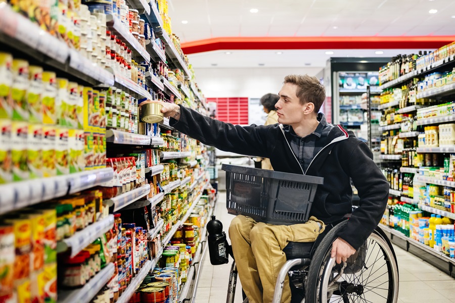 A man using a wheelchair shops in a grocery store, reaching for a can on a high shelf while holding a shopping basket in his lap.