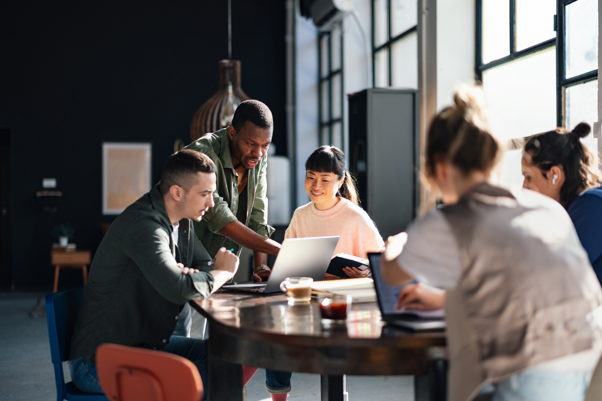 A diverse team collaborates around a table, reviewing ideas on a laptop in a bright, modern workspace, showcasing teamwork, creativity, and problem-solving.