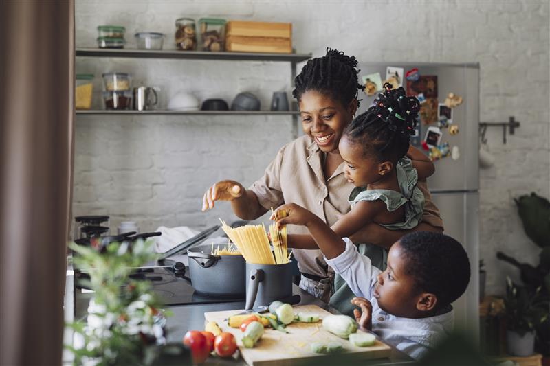 Mother and two children cooking together in a kitchen, adding spaghetti to a pot on the stove.
