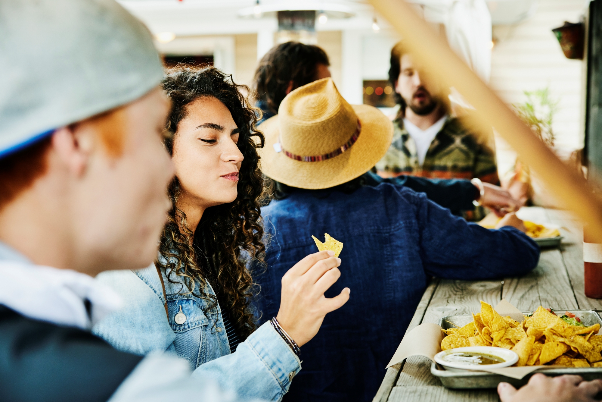 Group of young people sitting at a wooden table eating tortilla chips with salsa at an outdoor gathering.