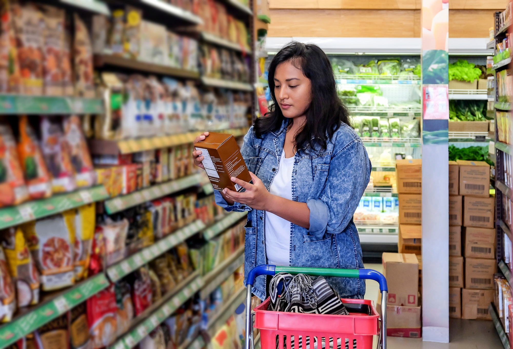 Woman in a denim jacket reading product packaging in a grocery store aisle with a shopping cart containing a bag.