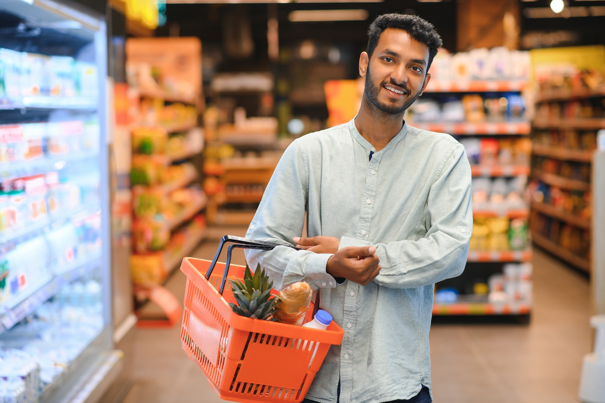 Smiling man in a light blue shirt holding an orange shopping basket with groceries in a supermarket aisle.