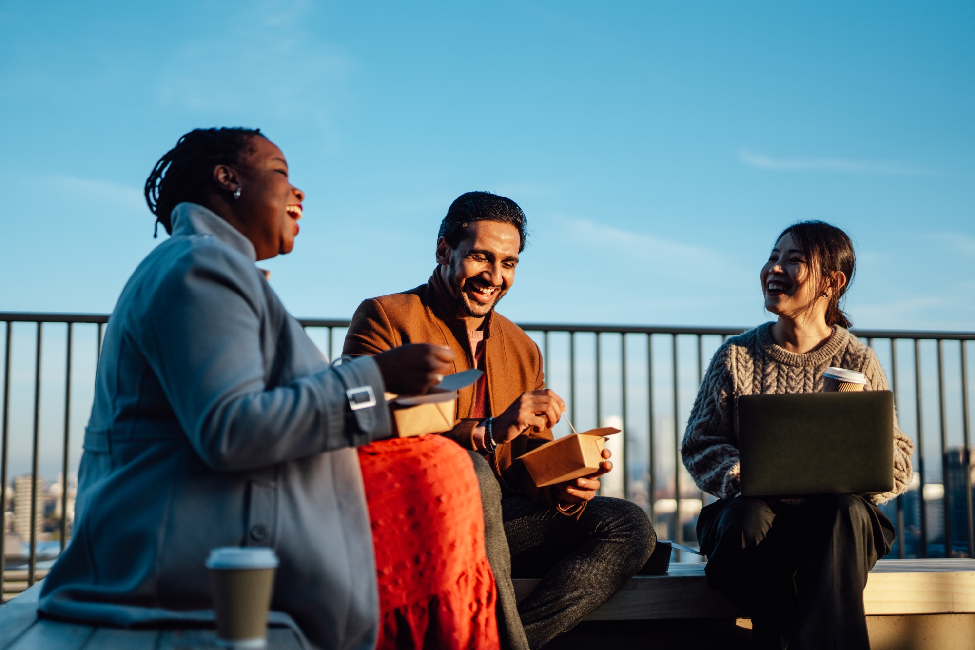 Three diverse colleagues laughing and eating lunch outdoors with a city view, one holding a laptop and coffee.