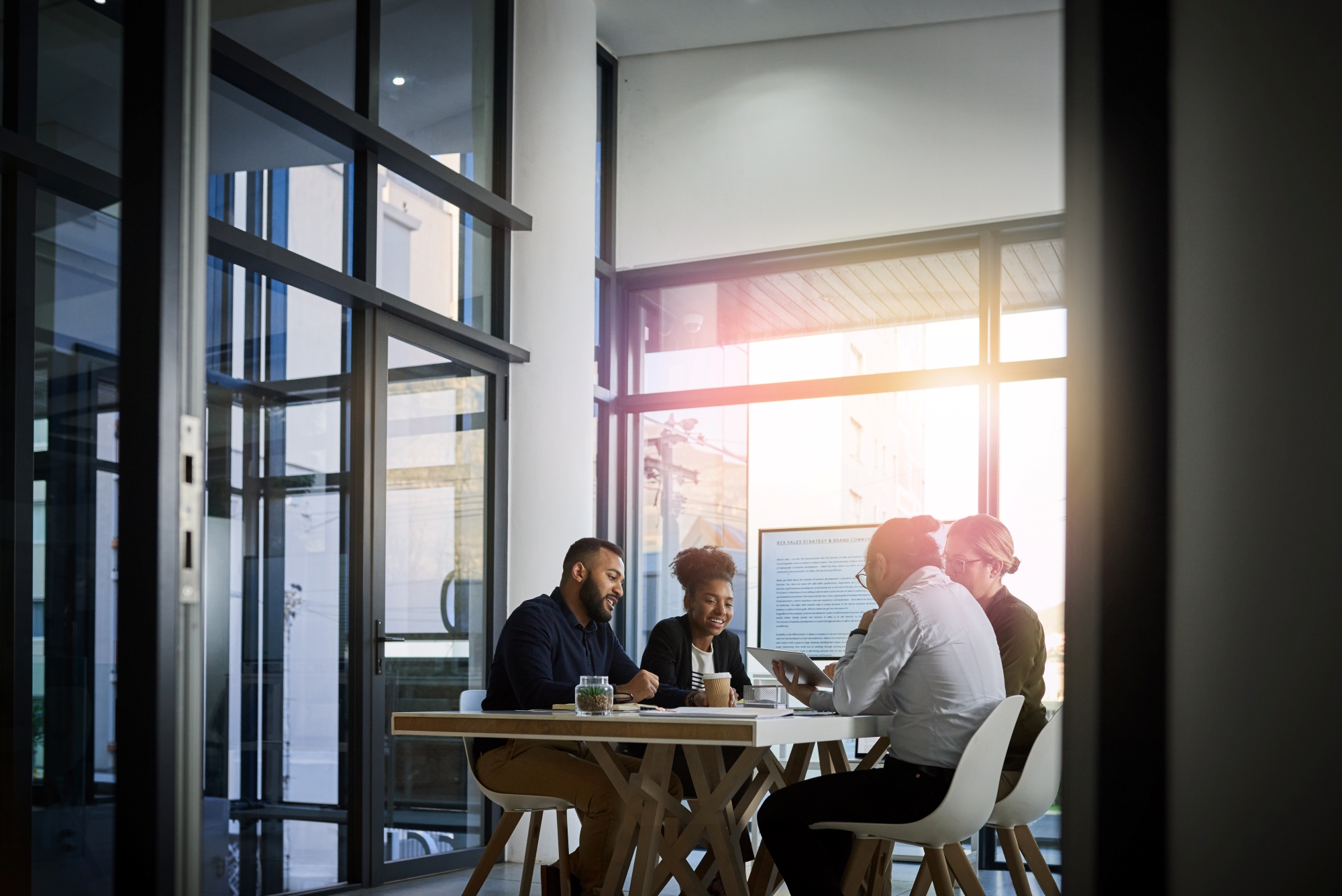 Four colleagues sitting around a table in a modern office with large windows, discussing and smiling while reviewing a tablet.