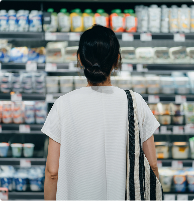 Woman with a striped tote bag standing and looking at refrigerated shelves in a grocery store.