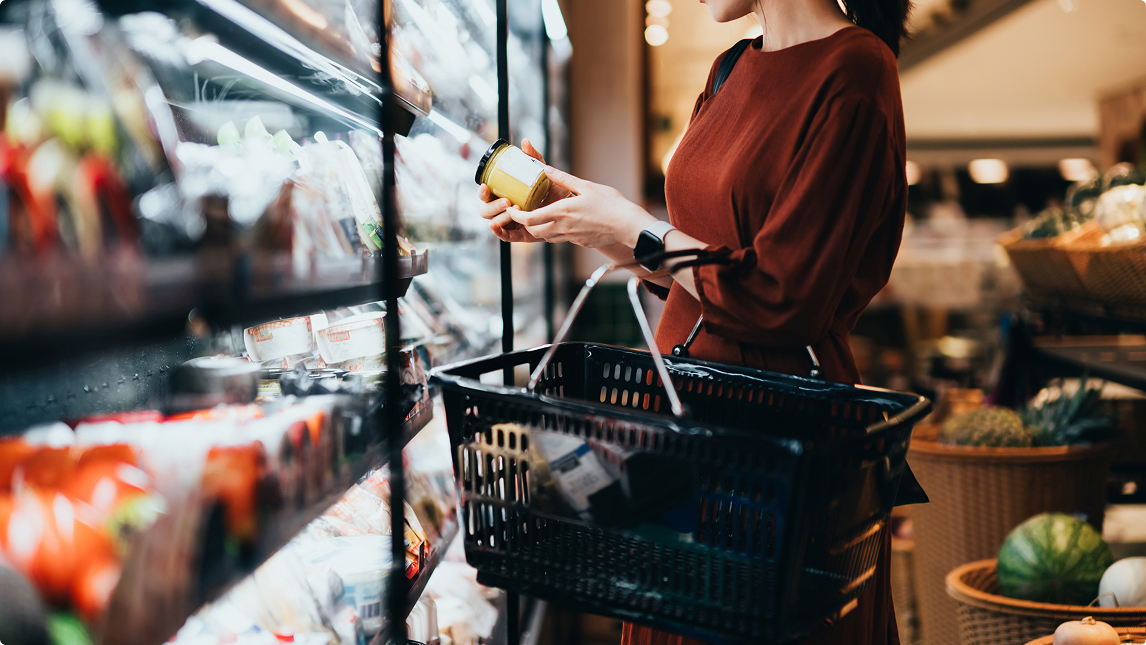 Woman in a red dress holding a jar and shopping with a basket in a grocery store aisle.