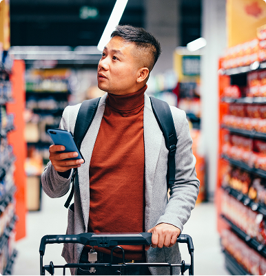 Man with a backpack holding a smartphone and pushing a shopping cart in a grocery store aisle.